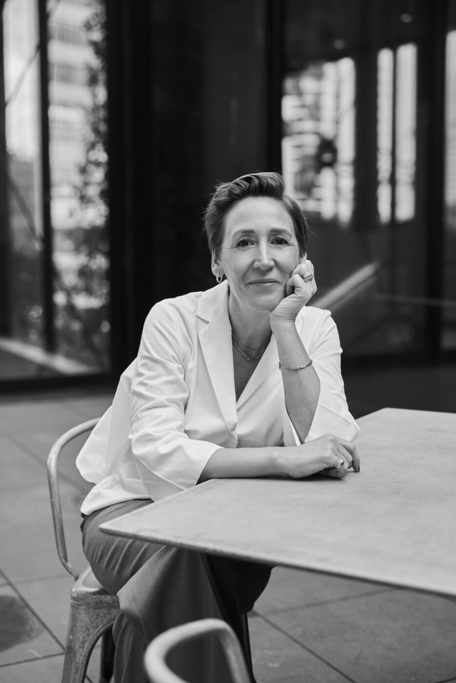 Black and white portrait of Alice Herald, seated at a table, wearing a white blazer and smiling in a modern, well-lit space