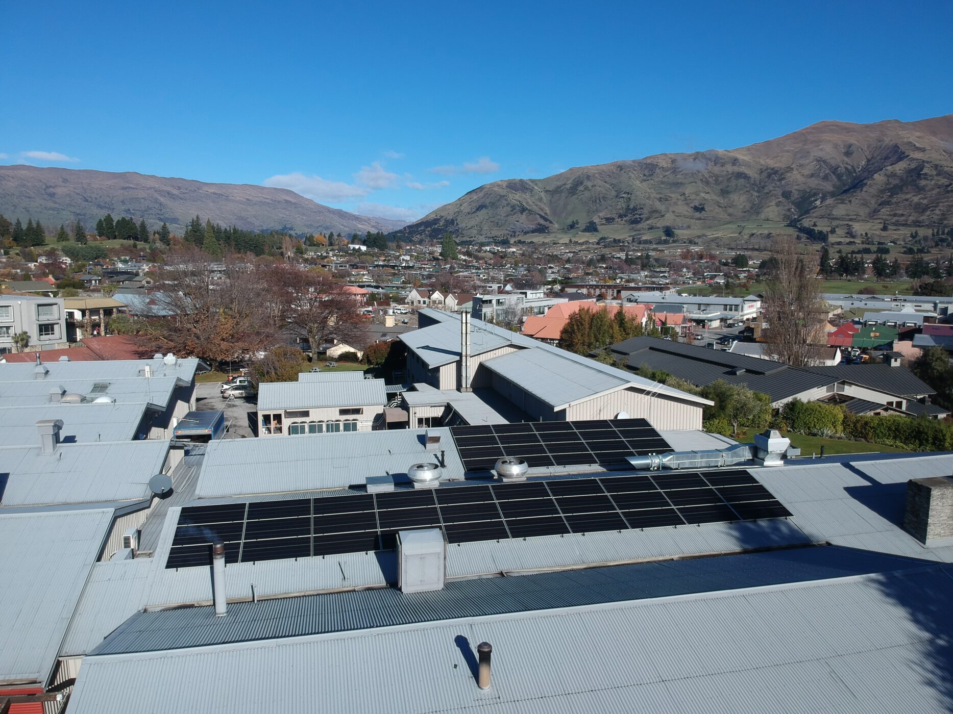 Aerial view of The Wānaka Hotel with solar panels installed across its rooftop, situated within Wānaka township surrounded by mountains